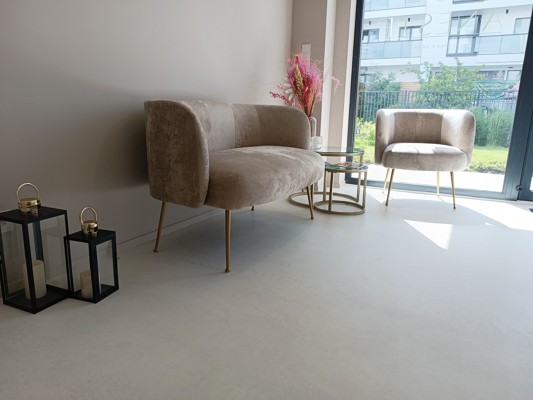 Jasnoszara, gładka posadzka w pomieszczeniu z beżową sofą i fotelem na złotych nóżkach, stolikami kawowymi i dekoracyjnymi lampionami; widok na zewnątrz przez duże okno.