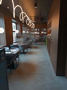 Wnętrze restauracji z szarą, gładką posadzką, nowoczesnym oświetleniem w postaci spiralnych lamp i ciemnymi meblami.