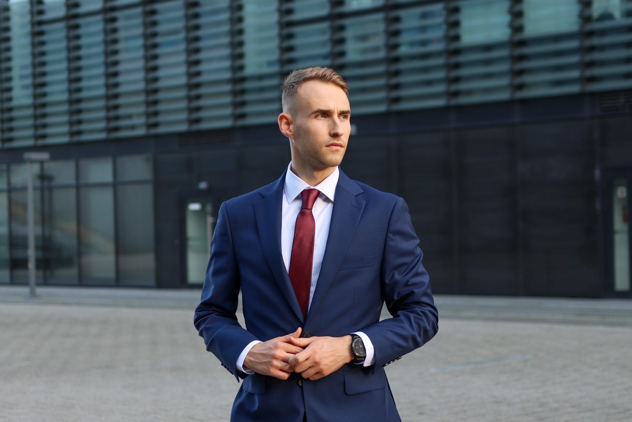 Młody mężczyzna w granatowym garniturze i bordowym krawacie, stoi na tle nowoczesnego budynku. Spojrzenie skierowane w bok, zegarek na nadgarstku.