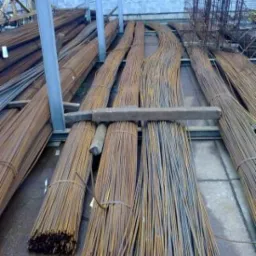 Sztaple wiązek prętów żebrowanych na metalowym regale w magazynie, widoczne ślady rdzy i zabrudzenia.