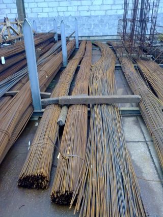 Sztaple wiązek prętów żebrowanych na metalowym regale w magazynie, widoczne ślady rdzy i zabrudzenia.