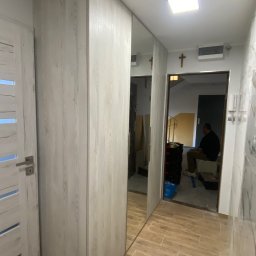 Meble na wymiar Tom - Nowoczesna szafa wnękowa z lustrzanymi drzwiami, imitująca drewno, w przedpokoju z płytkami podłogowymi w odcieniach brązu; w tle widoczny mężczyzna siedzący na klatce schodowej.