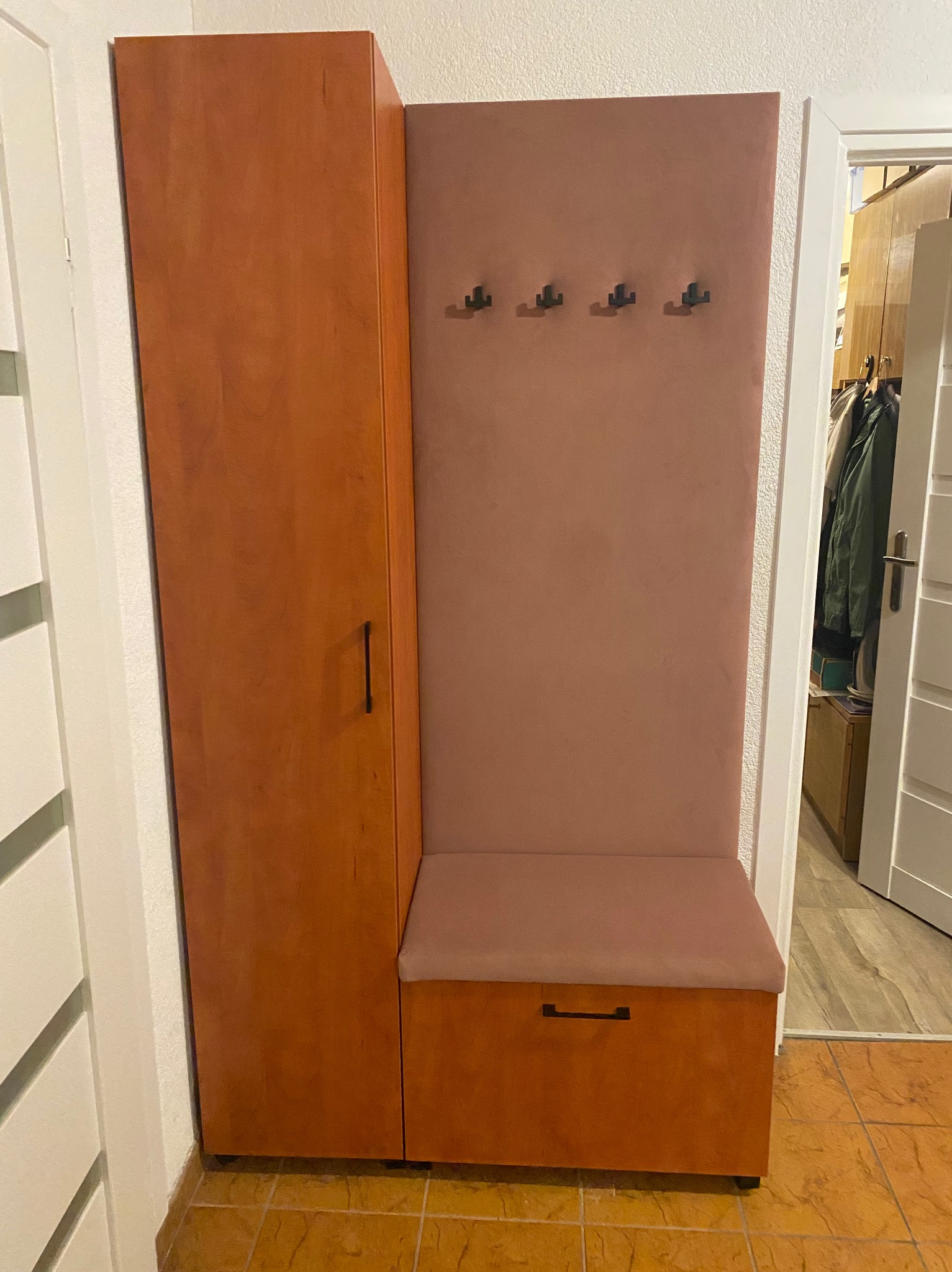 Komplet mebli do przedpokoju: wysoka szafa z pojedynczym drzwiami, panel z czterema wieszakami i siedziskiem z szufladą w kolorze ciepłego brązu i pudrowego różu, widoczne płytki podłogowe...