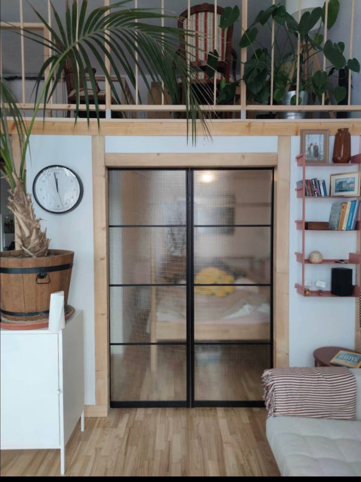 Dwuskrzydłowe drzwi szklane z czarnymi szprosami w drewnianej ościeżnicy, widok z salonu na sypialnię, na ścianie zegar i metalowy regał, podwieszona antresola z roślinami doniczkowymi.