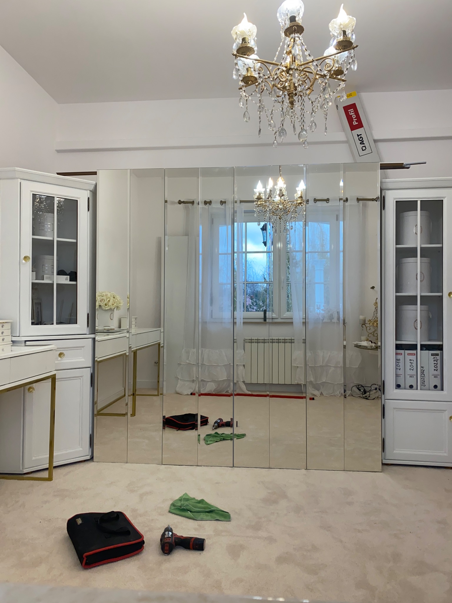 Montaż szafy wnękowej z lustrzanymi drzwiami przesuwnymi, widoczne narzędzia i akcesoria montażowe na podłodze, odbicie żyrandola w lustrach.