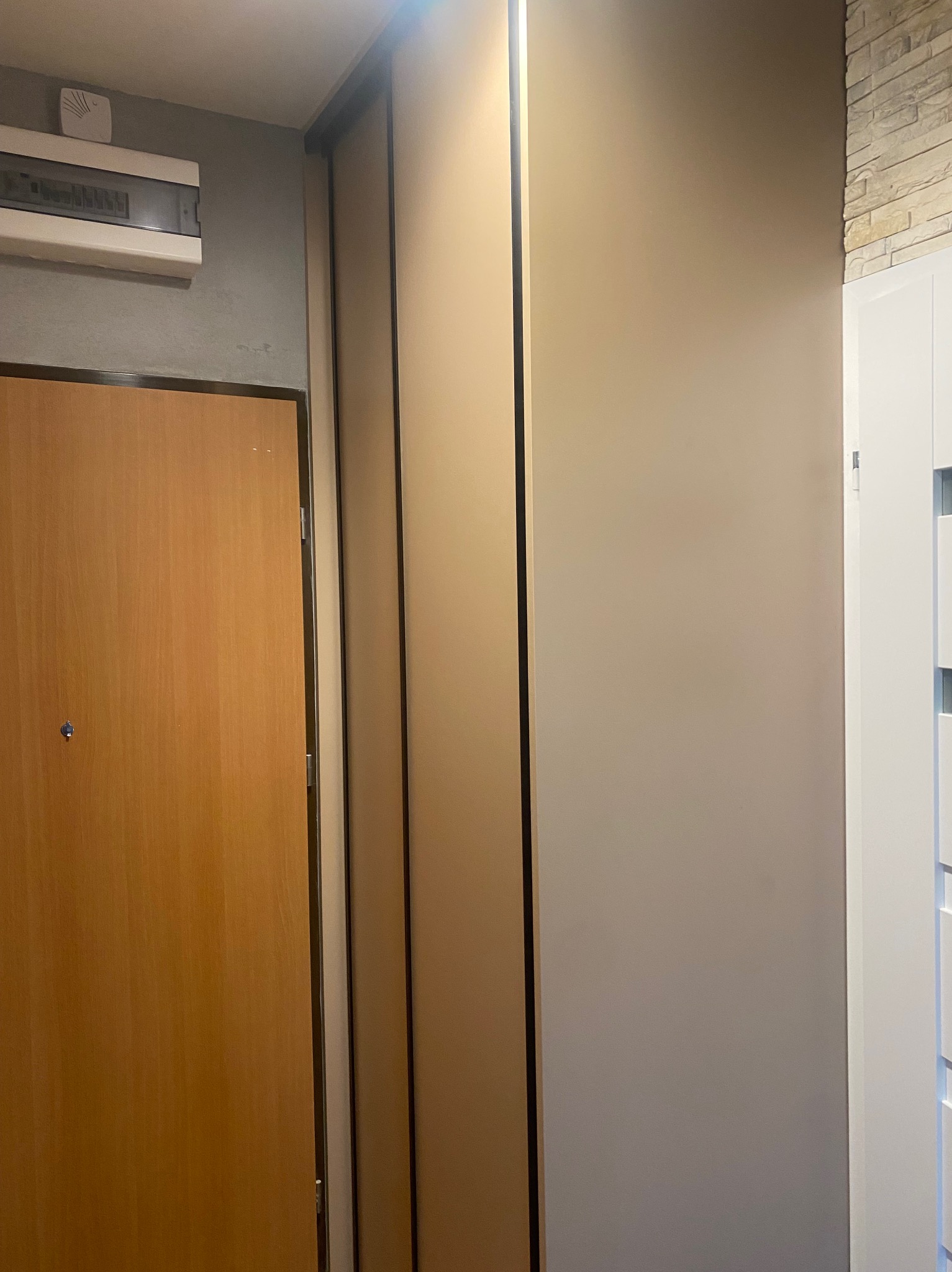 Wnętrze przedpokoju z zabudowaną szafą przesuwną w kolorze beżowym, drewnianymi drzwiami wejściowymi i fragmentem ściany wykończonej kamieniem dekoracyjnym.