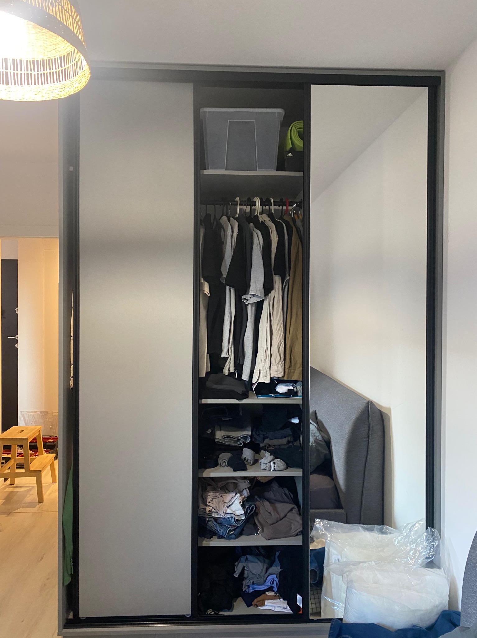 Wysoka szafa z przesuwnymi drzwiami, jedna strona z lustrem, widoczne ubrania wewnątrz, na podłodze folia zabezpieczająca, w tle fragment pokoju z krzesłem.