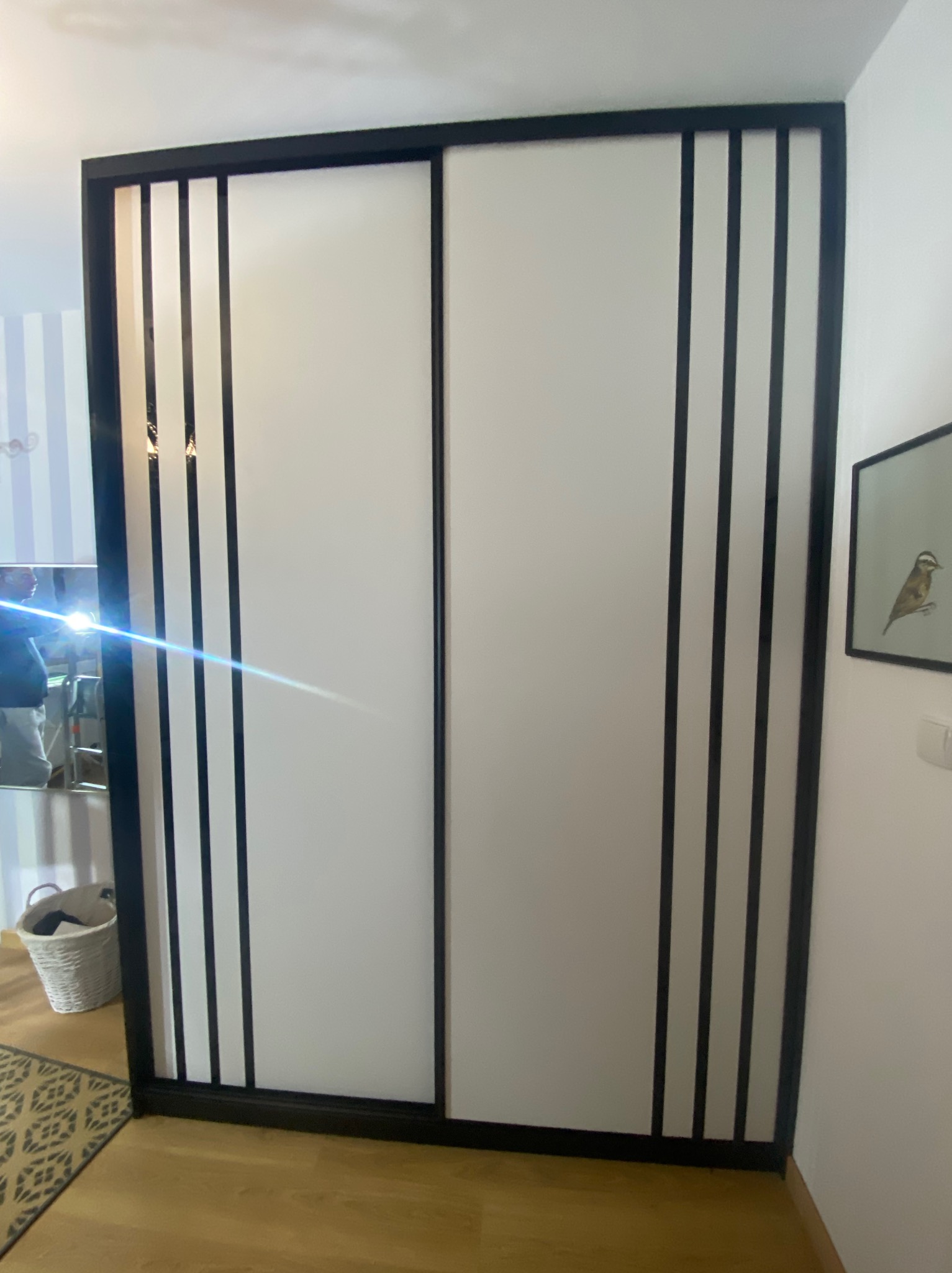 Dwudrzwiowa szafa przesuwna z białymi frontami i czarnymi, pionowymi listwami na obrzeżach, widoczna z bliskiej perspektywy w jasnym wnętrzu.