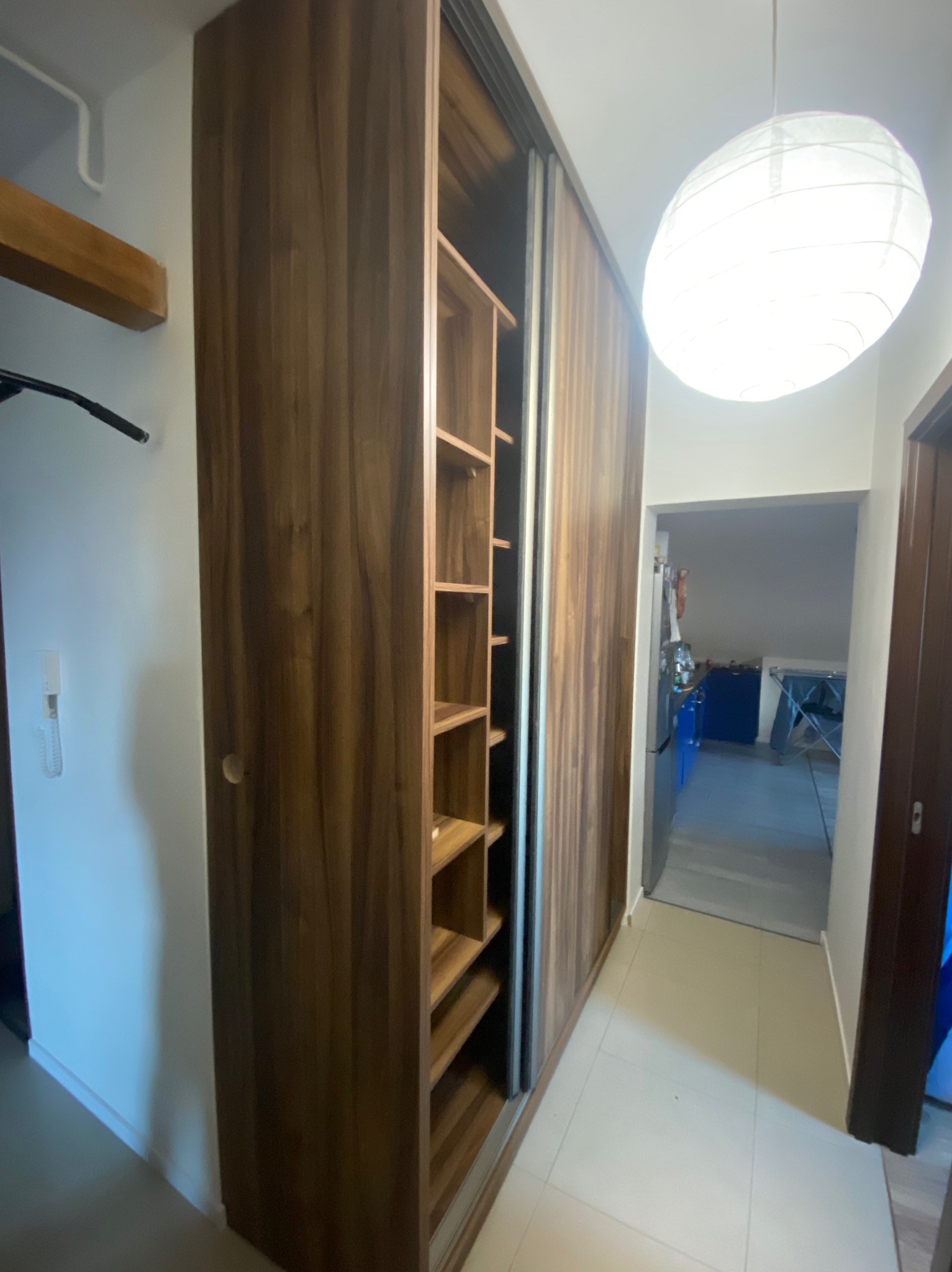 Wnętrze przedpokoju z szafą wnękową z przesuwnymi drzwiami z forniru w odcieniach brązu, częściowo otwartą z widocznymi półkami, oświetloną wiszącą lampą kulą.