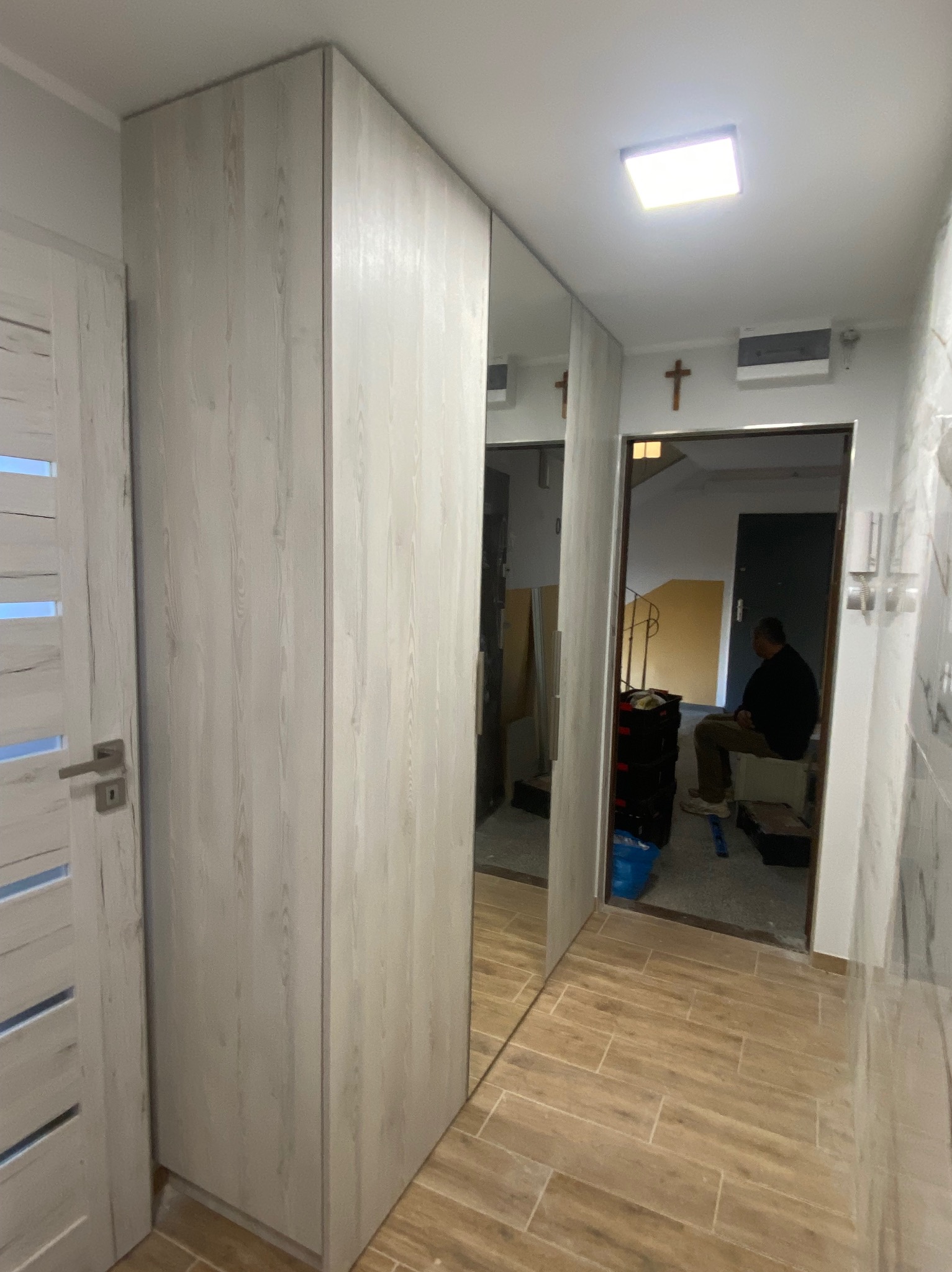 Nowoczesna szafa wnękowa z lustrzanymi drzwiami, imitująca drewno, w przedpokoju z płytkami podłogowymi w odcieniach brązu; w tle widoczny mężczyzna siedzący na klatce schodowej.