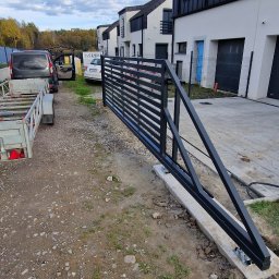 LOFT WELDING Krystian Kudela - Nowoczesna, czarna brama przesuwna z metalowymi, poziomymi szczeblami, zamontowana na betonowej podstawie. W tle domy w zabudowie szeregowej i błękitne niebo.