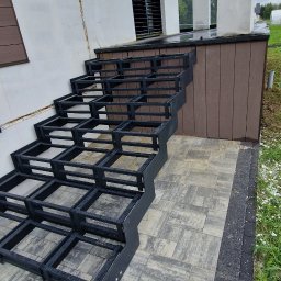 LOFT WELDING Krystian Kudela - Surowa konstrukcja schodów metalowych na zewnątrz budynku, z widoczną ramą i stopniami, czekająca na wykończenie. Szara kostka brukowa i fragment elewacji w tle.