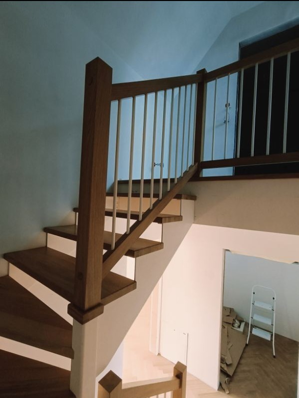 Drewniane schody z białą balustradą, widok z dołu, w tle widoczne schody i resztki materiałów budowlanych.