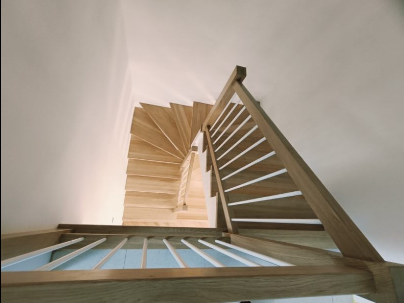 Widok z góry na spiralne drewniane schody z jasnymi, pionowymi listwami balustrady i białymi ścianami, oświetlone naturalnym światłem.