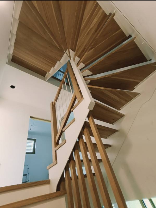 Spiralne schody z drewnianymi stopniami i białymi balustradami, widok z dołu, z oknem i drabiną widoczną w tle.