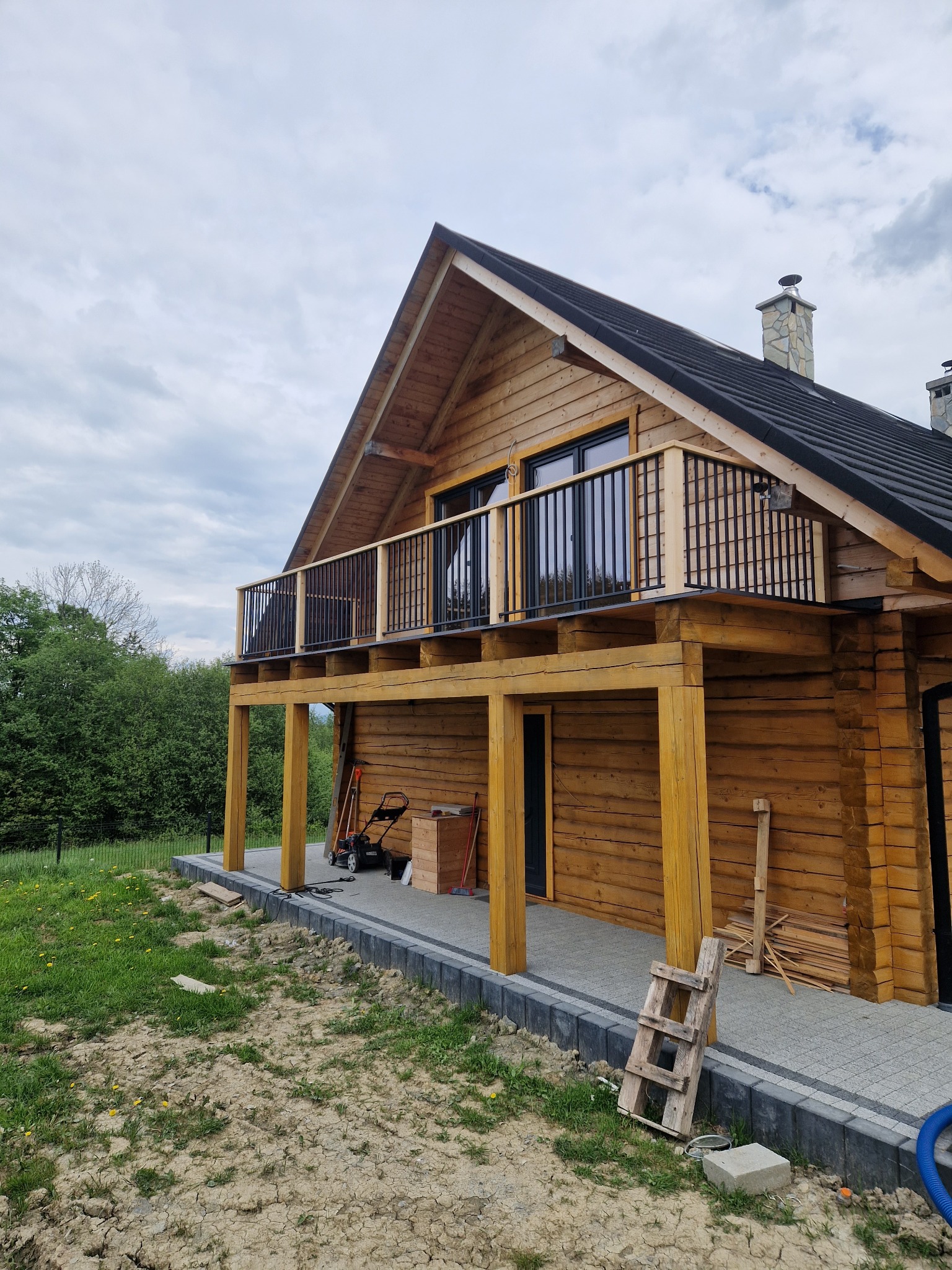 Drewniany dom z balkonem i czarną, metalową balustradą, widok z zewnątrz, częściowo zagospodarowany teren wokół budynku, drabina oparta o ścianę.
