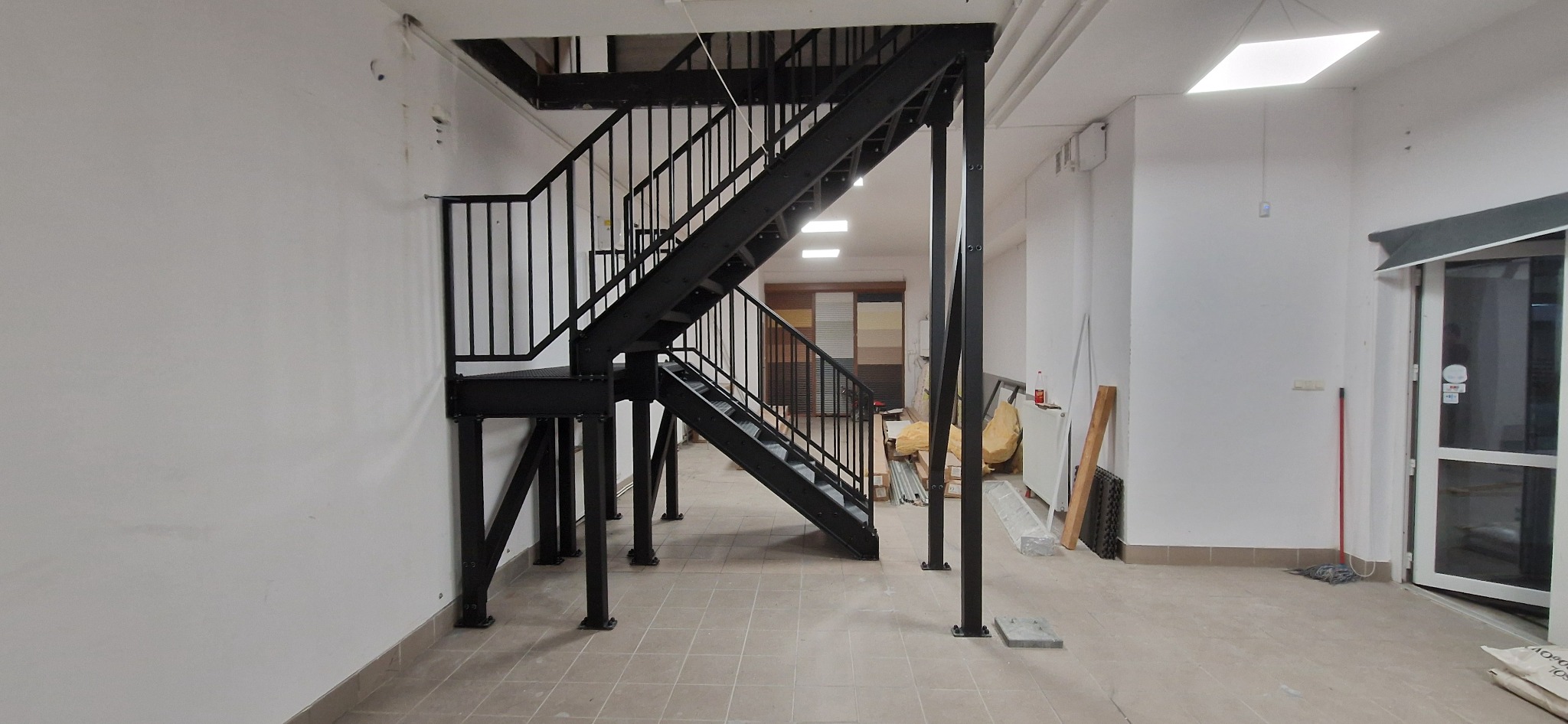 Czarne schody metalowe z balustradą w jasnym wnętrzu. Widoczne mocowania do ściany i podłogi. Ujęcie z dołu, ukazujące konstrukcję schodów.