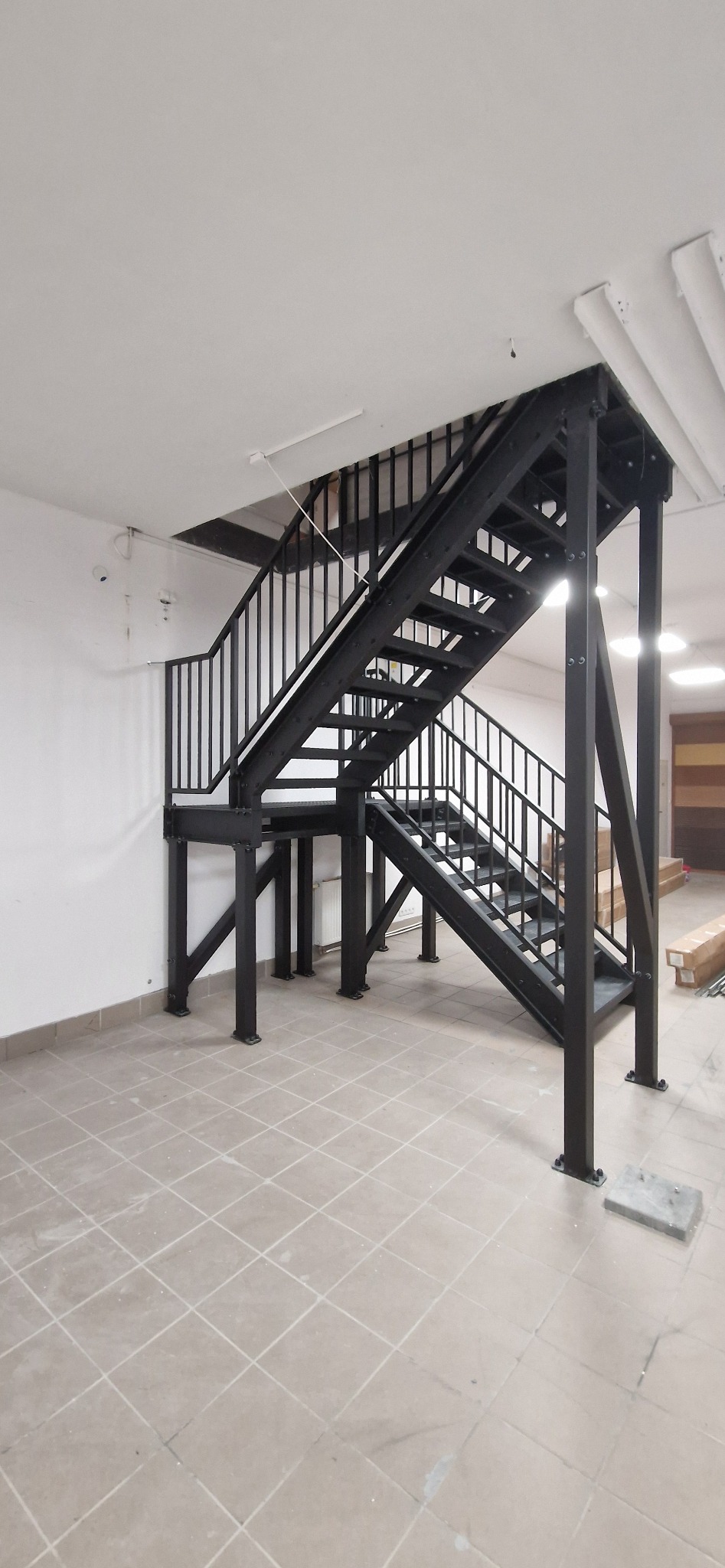 Dwubiegowe schody metalowe w czarnym kolorze, z prostą balustradą, zamontowane w jasnym pomieszczeniu z płytkami podłogowymi. Widok z dołu.