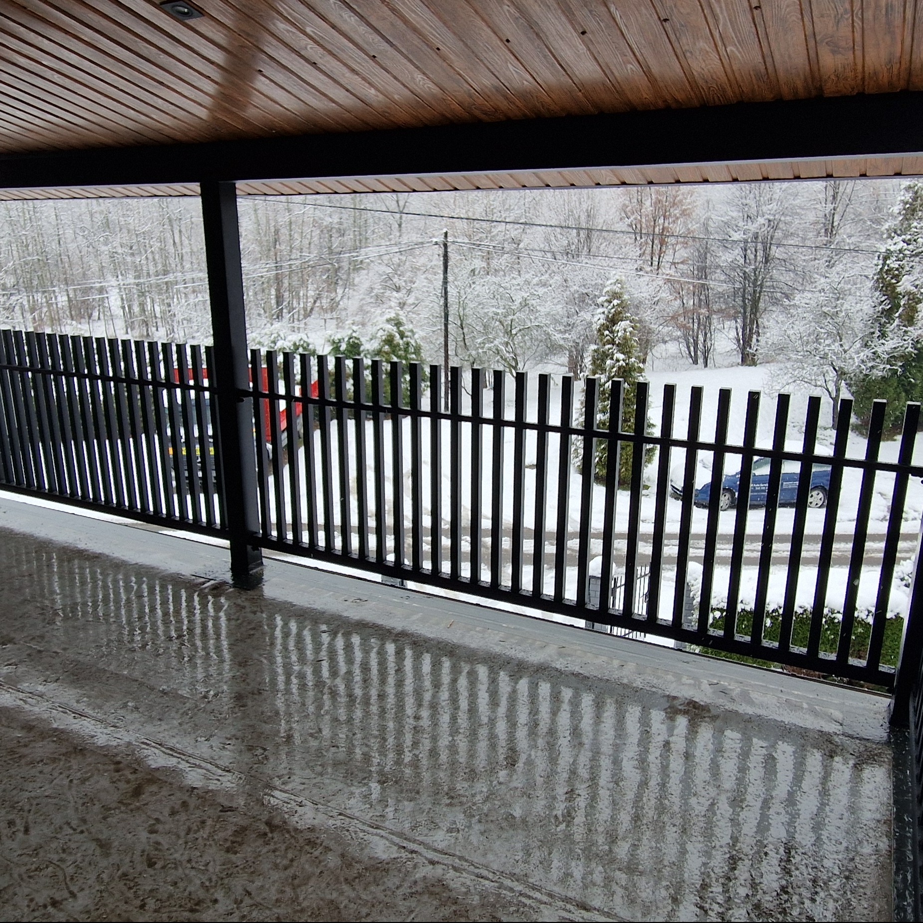 Czarna, metalowa balustrada balkonu z widokiem na zimowy krajobraz. Na podłodze balkonu widoczne odbicie balustrady. Drewniane wykończenie sufitu.
