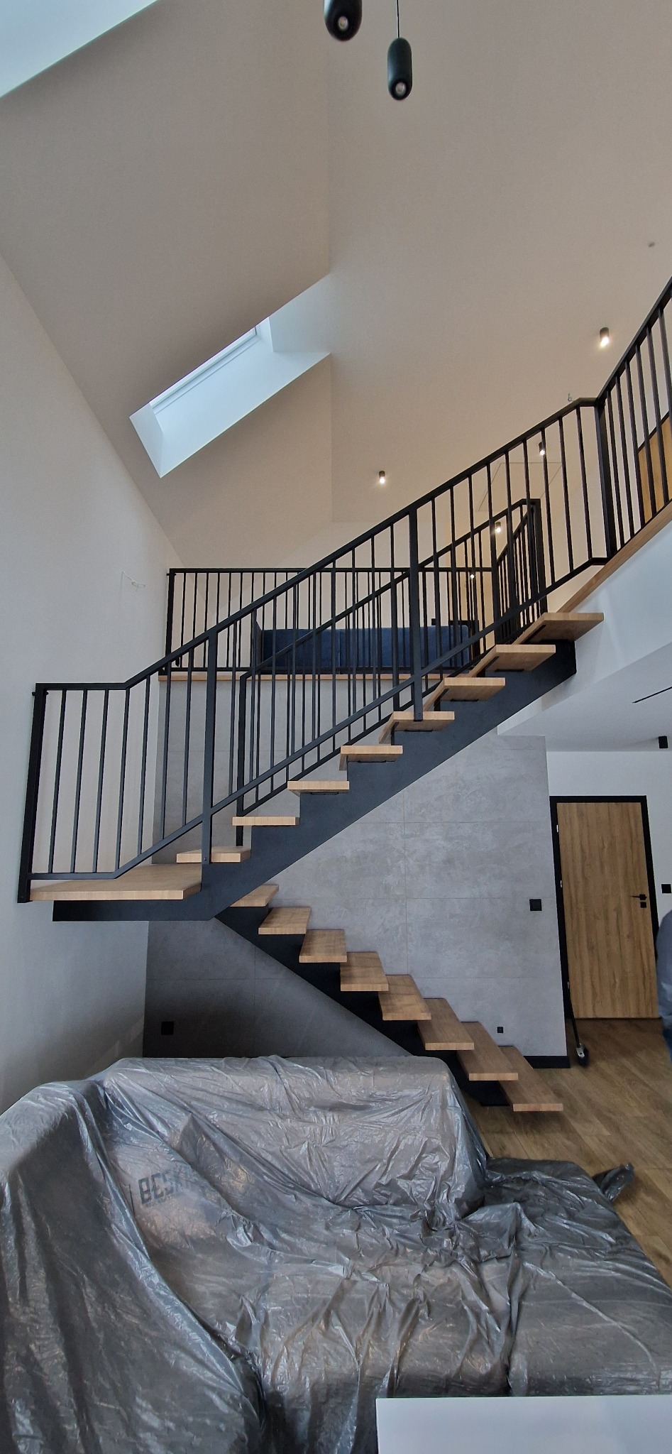 Nowoczesne schody z drewnianymi stopniami i czarną metalową balustradą w jasnym wnętrzu z oknem dachowym. Sofa zabezpieczona folią ochronną.