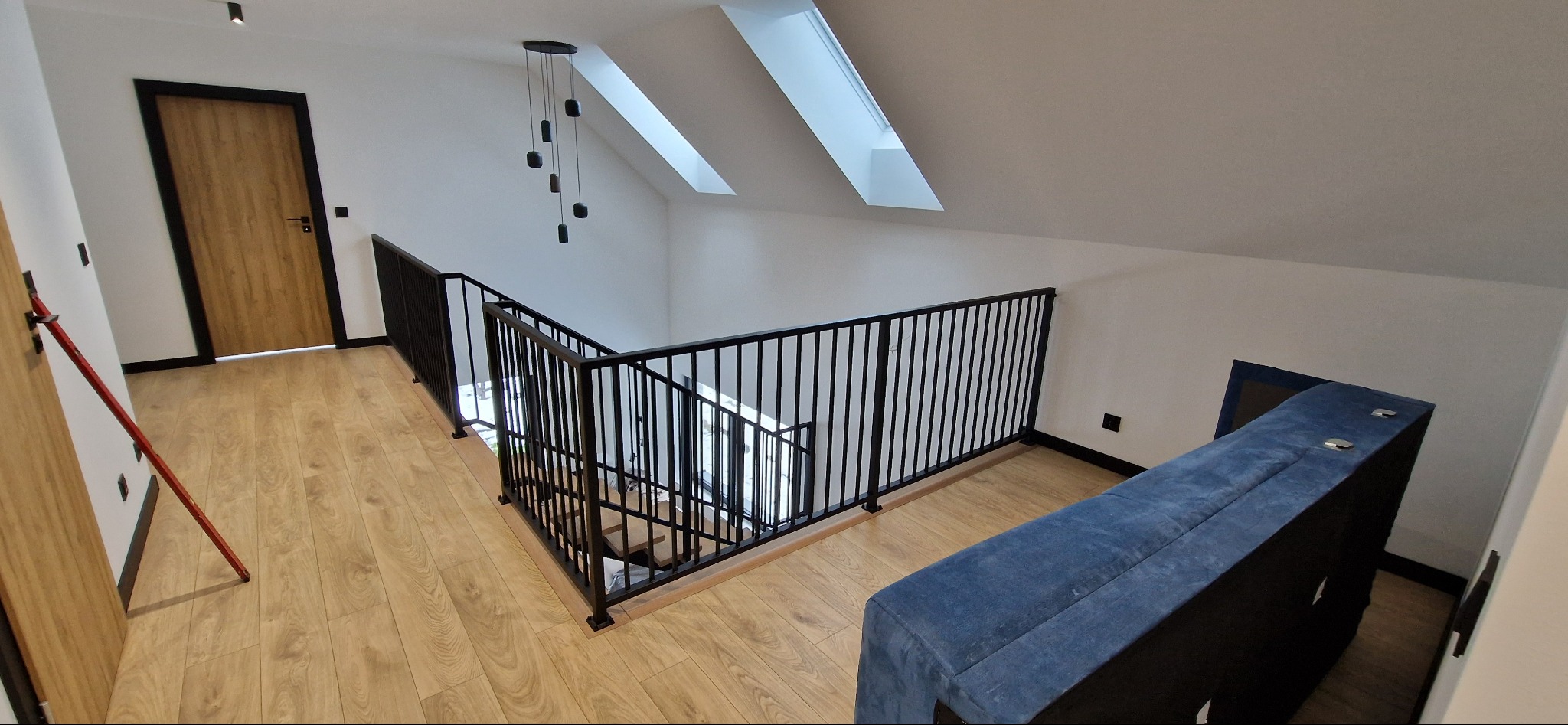 Nowoczesne wnętrze z czarną, metalową balustradą schodową na antresoli. Widoczne schody, drewniana podłoga i okna dachowe. Minimalistyczny design.