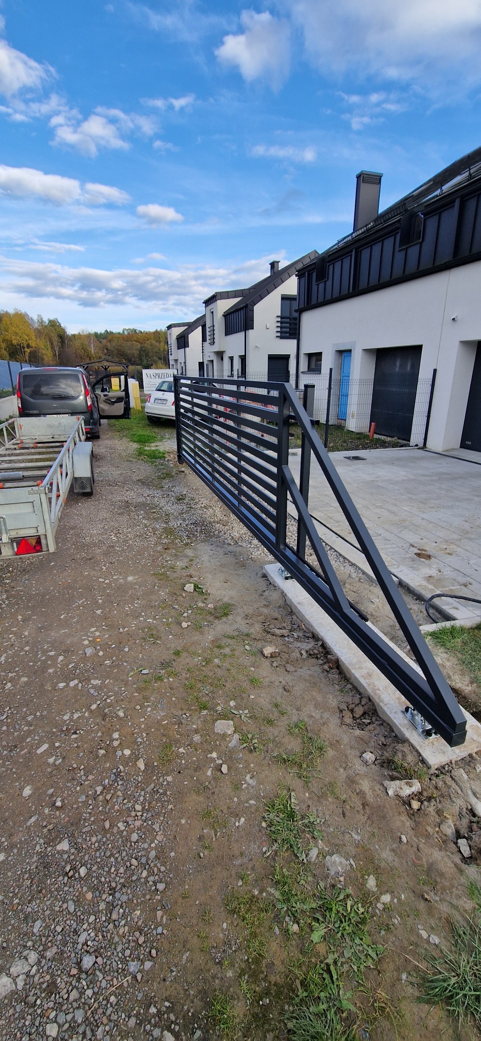Nowoczesna, czarna brama przesuwna z metalowymi, poziomymi szczeblami, zamontowana na betonowej podstawie. W tle domy w zabudowie szeregowej i błękitne niebo.