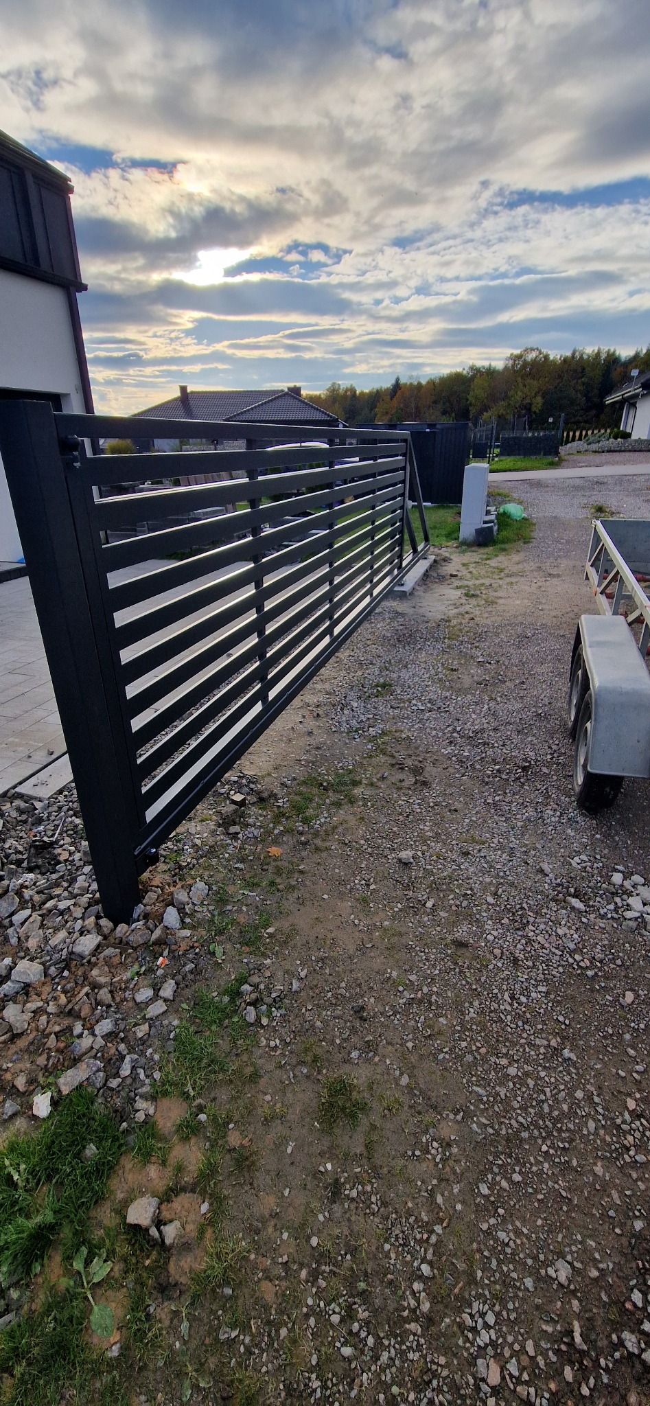 Nowoczesna, pozioma, czarna brama metalowa z szerokimi szczeblami, zamontowana przy posesji. Widoczne elementy konstrukcyjne i otoczenie posesji.