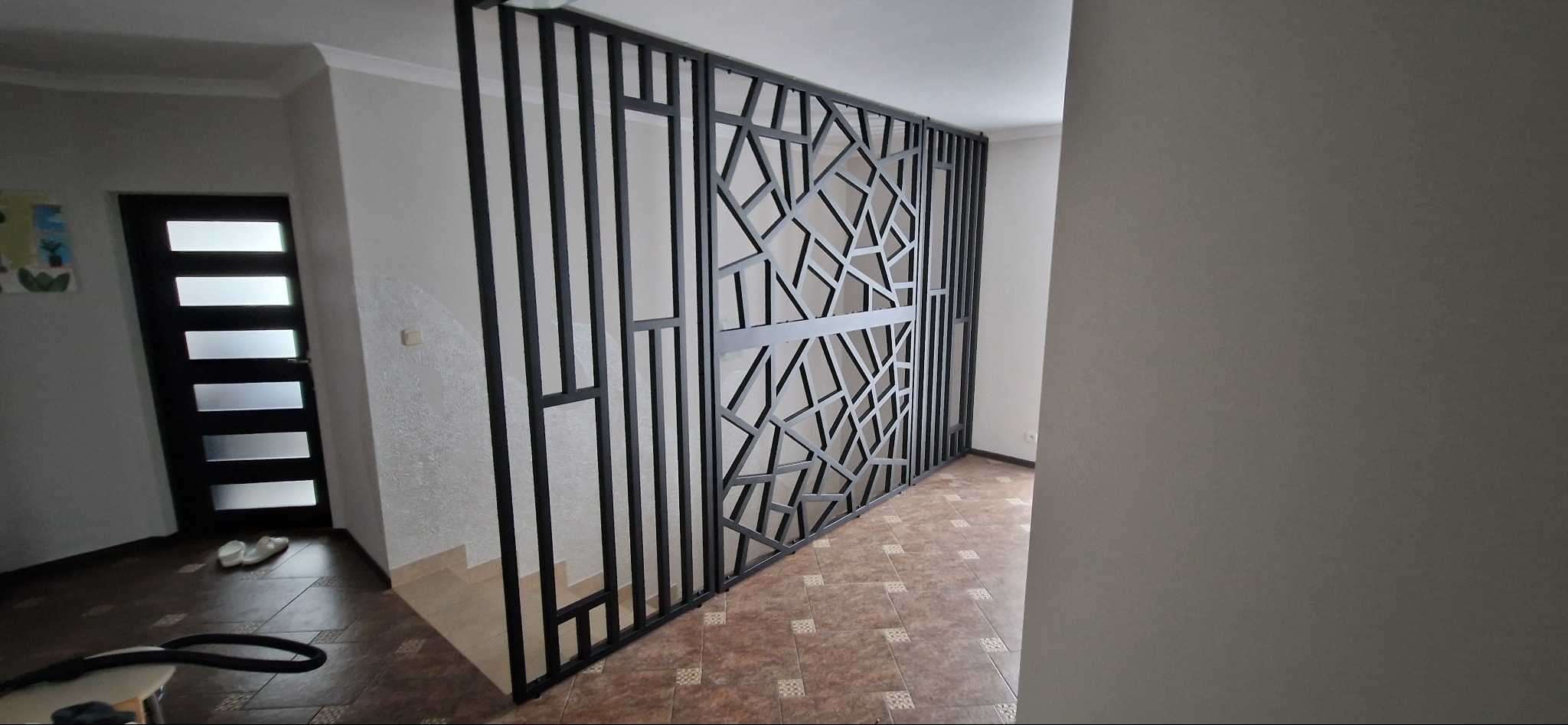 Nowoczesna, czarna ścianka działowa z metalu z geometrycznym wzorem, oddzielająca przestrzeń w jasnym wnętrzu. Widoczne schody i drzwi w tle.