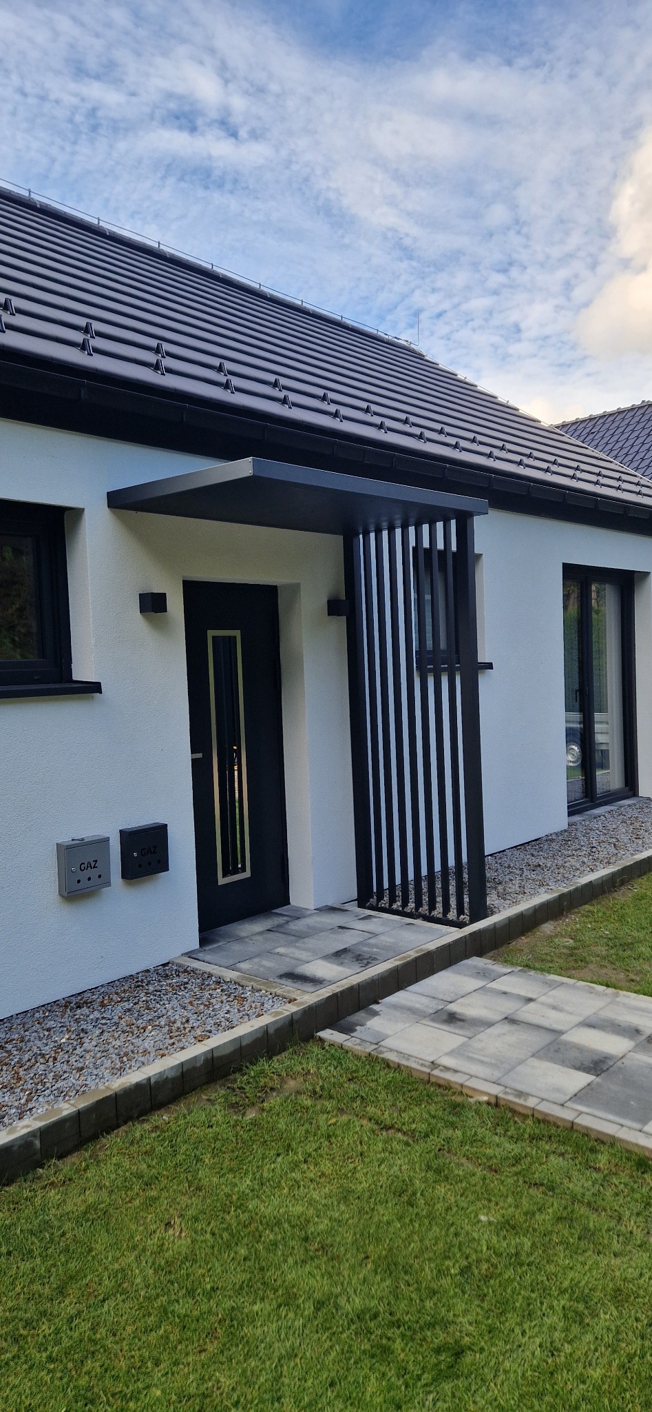 Nowoczesne zadaszenie nad wejściem do domu z czarnymi, pionowymi elementami dekoracyjnymi. Jasna elewacja, szara kostka brukowa i trawnik.