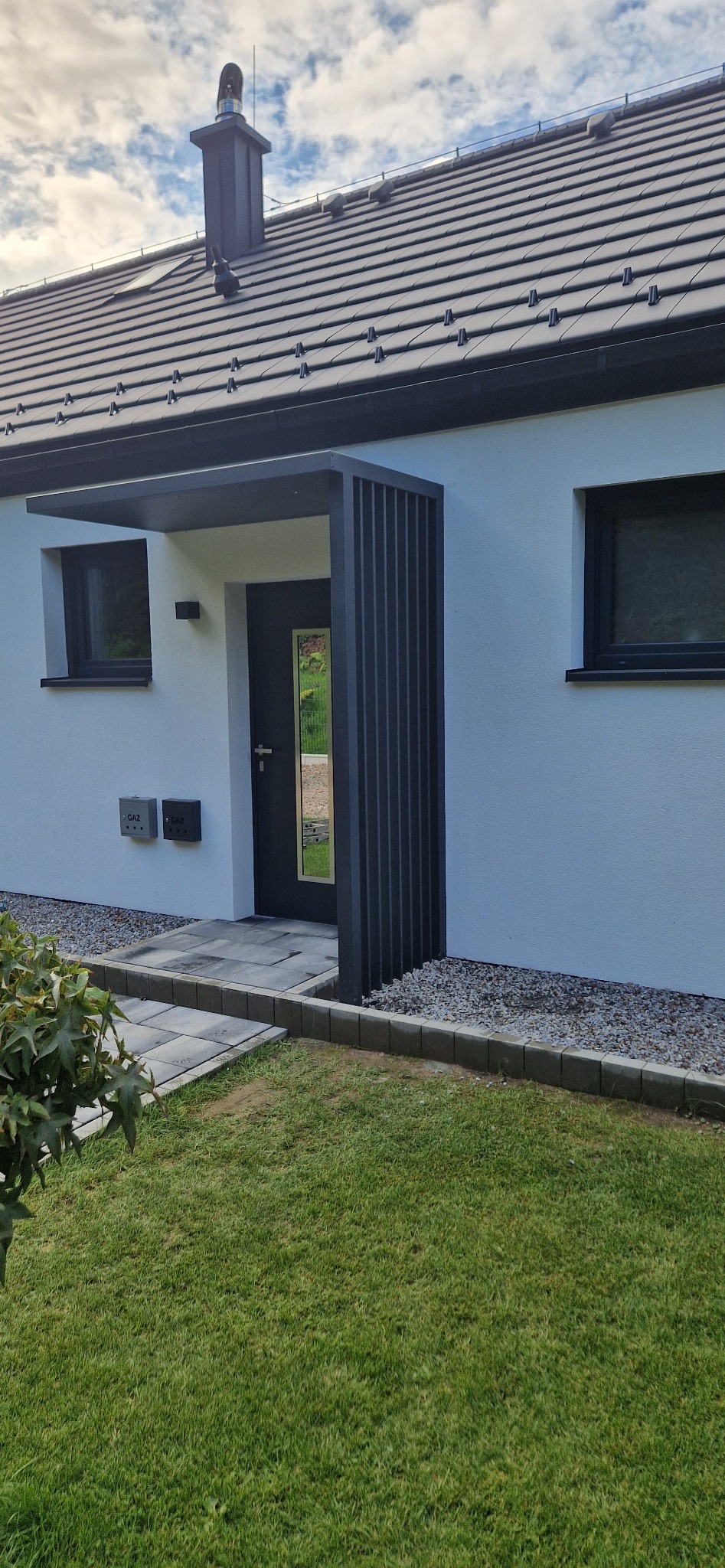 Nowoczesne zadaszenie nad wejściem do domu z pionowymi, metalowymi elementami. Minimalistyczny design, szara kolorystyka, kostka brukowa i trawnik przed budynkiem.