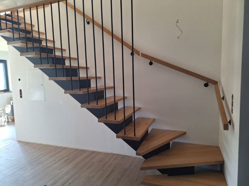 Nowoczesne schody z drewnianymi stopniami i czarnymi metalowymi wspornikami oraz poręczą z jasnego drewna, zamontowaną na białej ścianie.