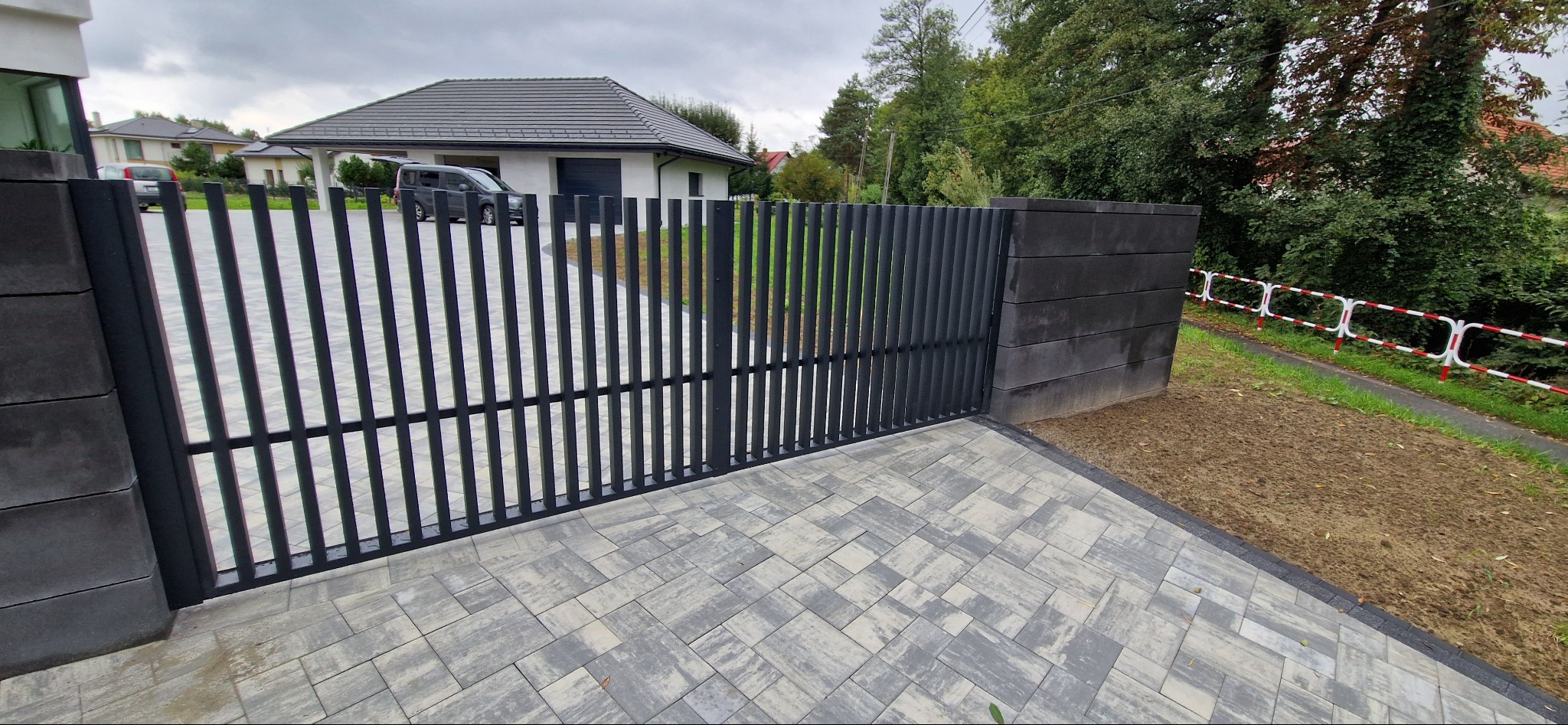 Nowoczesna, czarna brama posesyjna z pionowych profili, osadzona na betonowych słupkach. Kostka brukowa na podjeździe, w tle dom z szarą dachówką i zaparkowany samochód.