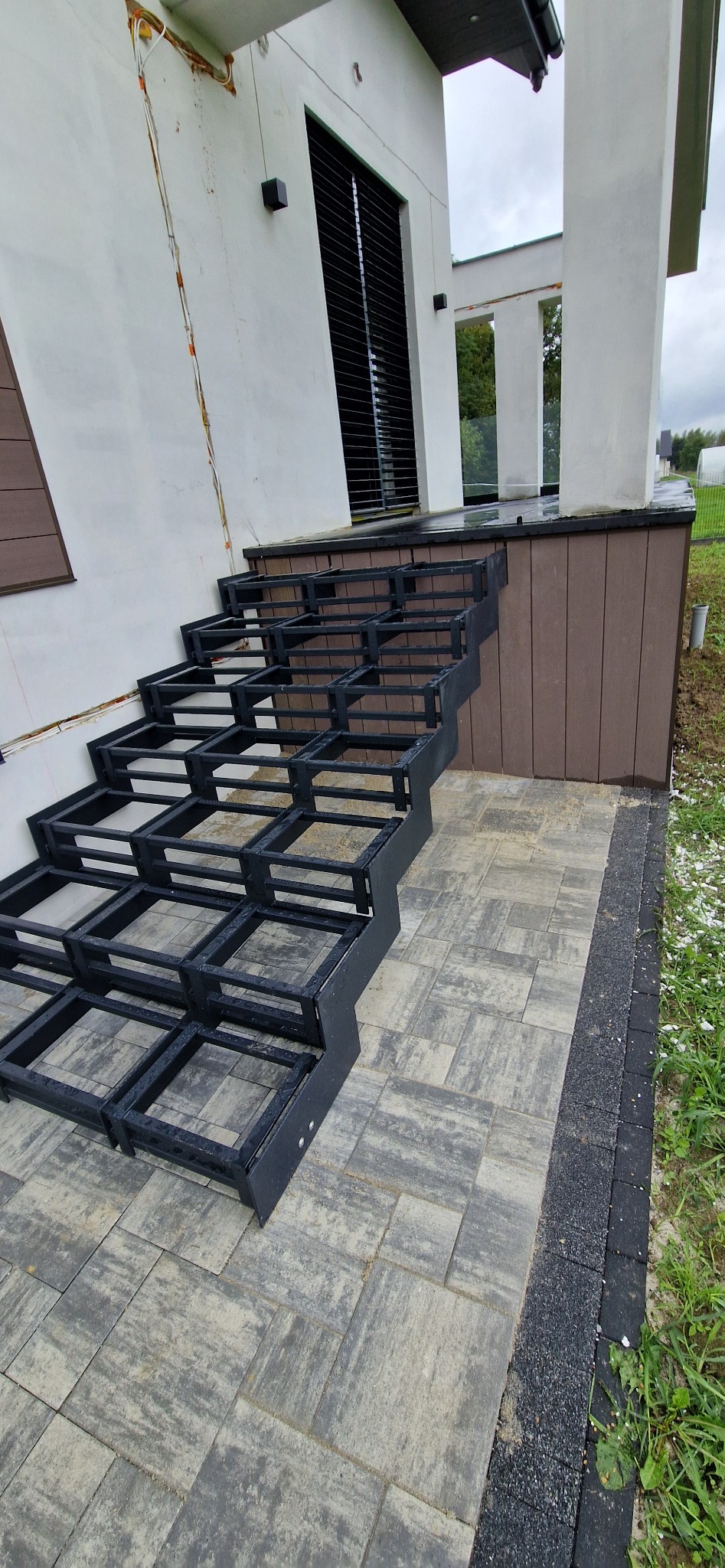 Surowa konstrukcja schodów metalowych na zewnątrz budynku, z widoczną ramą i stopniami, czekająca na wykończenie. Szara kostka brukowa i fragment elewacji w tle.