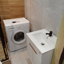 I.D.A.N BUD - Umywalka z szafką w białym kolorze i czarną armaturą, obok pralka Beko z wkrętakiem na górze, płytki imitujące drewno i marmur, widok z dołu.