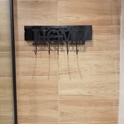 I.D.A.N BUD - Ściana wyłożona płytkami imitującymi drewno w Pabianicach z zamontowanym czarnym, metalowym wieszakiem na klucze z napisem 'HOME'.