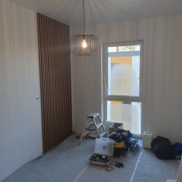 I.D.A.N BUD - Pokój w trakcie prac wykończeniowych: pionowe panele dekoracyjne z drewna na ścianie, podłoga zabezpieczona folią, widoczne narzędzia i drabina, okno częściowo zaklejone folią malarską, nowoczesna...