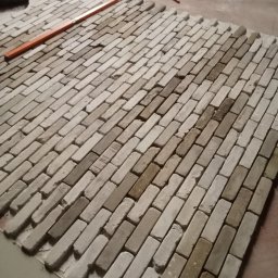 I.D.A.N BUD - Układanie cegieł klinkierowych na podłodze, widoczna poziomica i młotek, etap prac wykończeniowych.