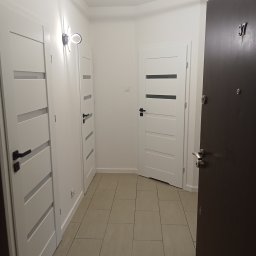 I.D.A.N BUD - Korytarz po remoncie z trzema białymi drzwiami z poziomymi przeszkleniami, podłoga wyłożona płytkami w odcieniach beżu, na ścianie nowoczesny kinkiet, widoczny numer mieszkania 37 na ciemnych...