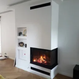 Nowoczesny kominek z czarnym obramowaniem wbudowany w białą ścianę z półkami, na podłodze parkiet ułożony w jodełkę francuską, widoczne płomienie w palenisku.