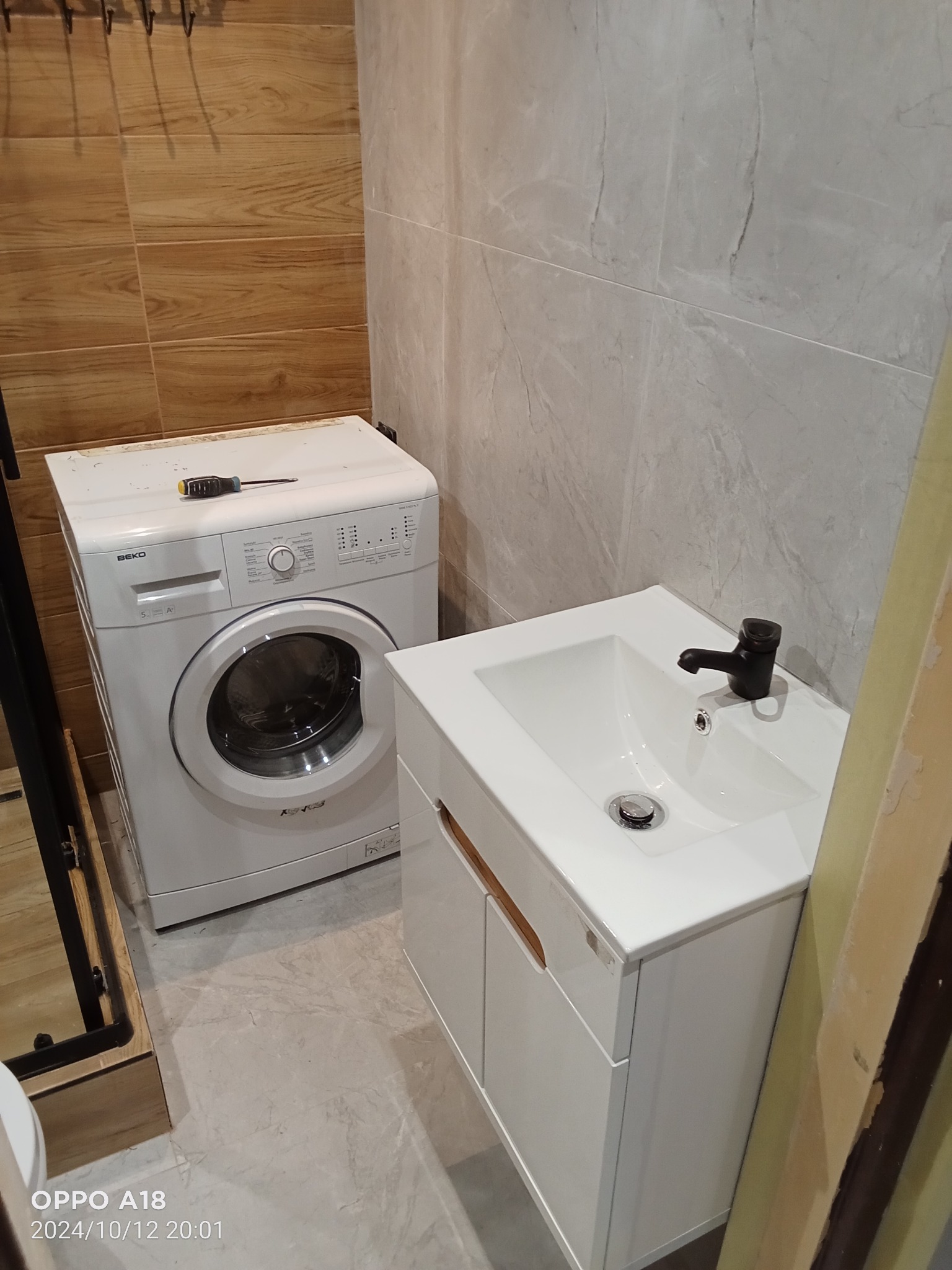 Umywalka z szafką w białym kolorze i czarną armaturą, obok pralka Beko z wkrętakiem na górze, płytki imitujące drewno i marmur, widok z dołu.