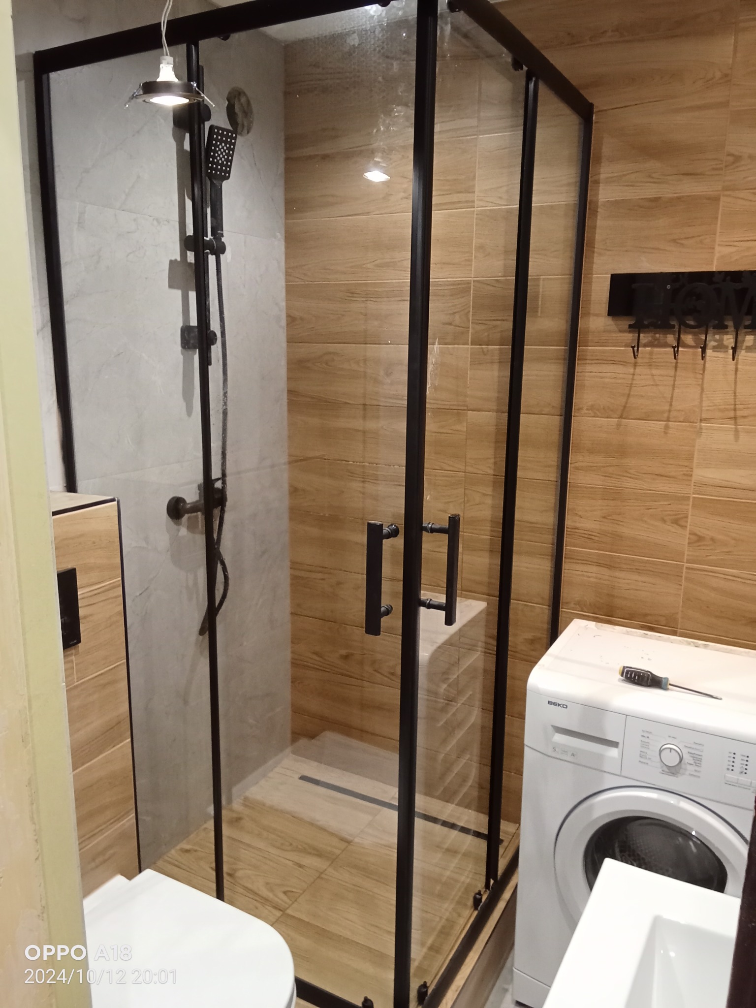 Nowoczesna kabina prysznicowa z czarnymi profilami, drewnopodobne płytki na ścianach, obok pralka, widoczny fragment umywalki i czarny wieszak na ręczniki.