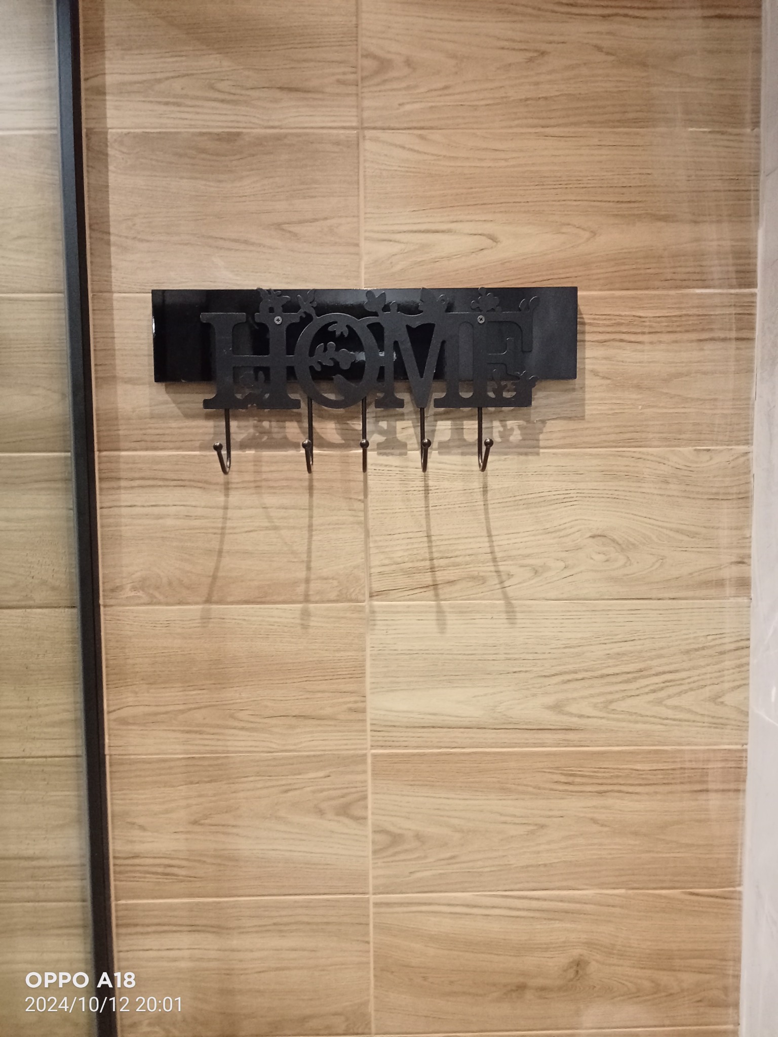 Ściana wyłożona płytkami imitującymi drewno w Pabianicach z zamontowanym czarnym, metalowym wieszakiem na klucze z napisem 'HOME'.