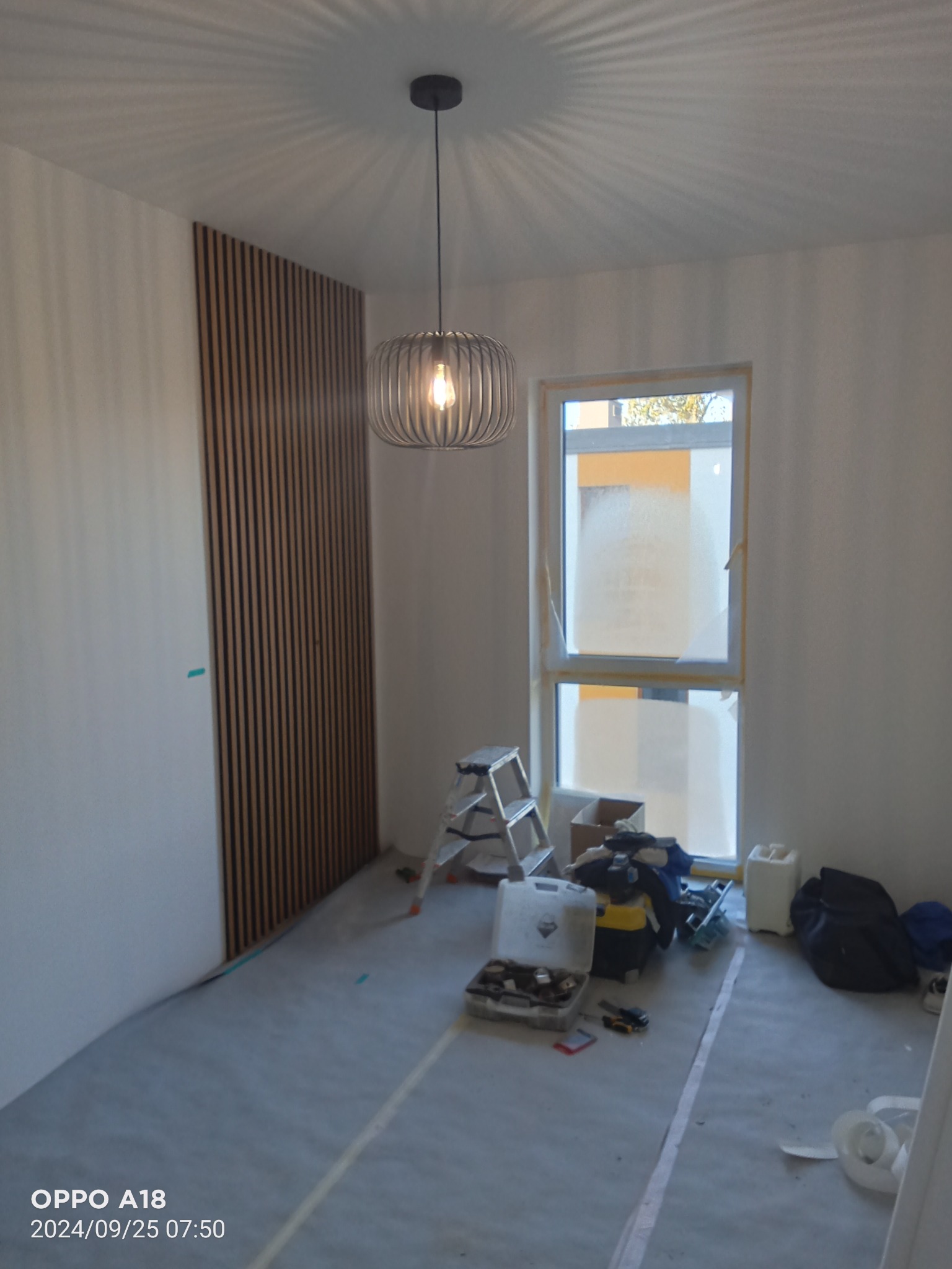 Pokój w trakcie prac wykończeniowych: pionowe panele dekoracyjne z drewna na ścianie, podłoga zabezpieczona folią, widoczne narzędzia i drabina, okno częściowo zaklejone folią malarską, nowoczesna...