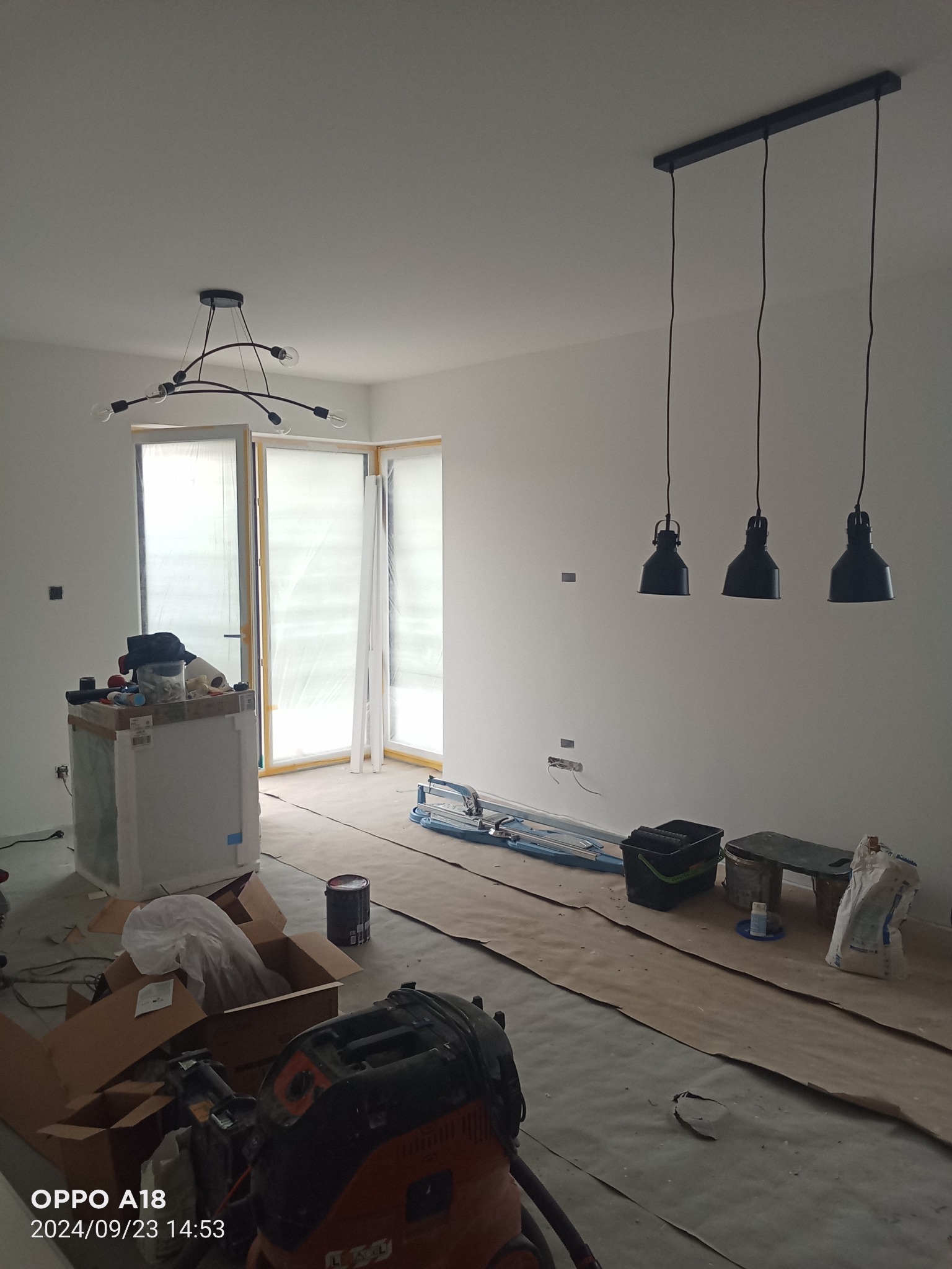 Wnętrze w trakcie remontu: widoczne zabezpieczone okna, surowe ściany z instalacją elektryczną, podłoga zabezpieczona kartonem, narzędzia i materiały budowlane, w tle lampy sufitowe.