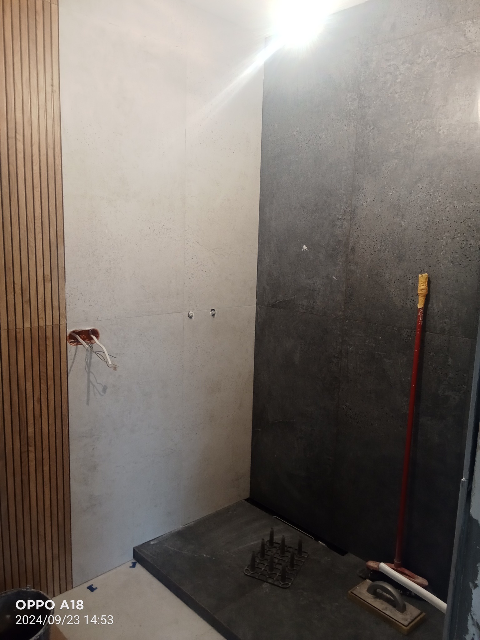 Wnętrze łazienki w trakcie remontu, widoczne płytki w odcieniach szarości i beżu, drewniane panele ścienne, wystające przewody elektryczne oraz narzędzia glazurnicze na podłodze, w tym paca i listwa.