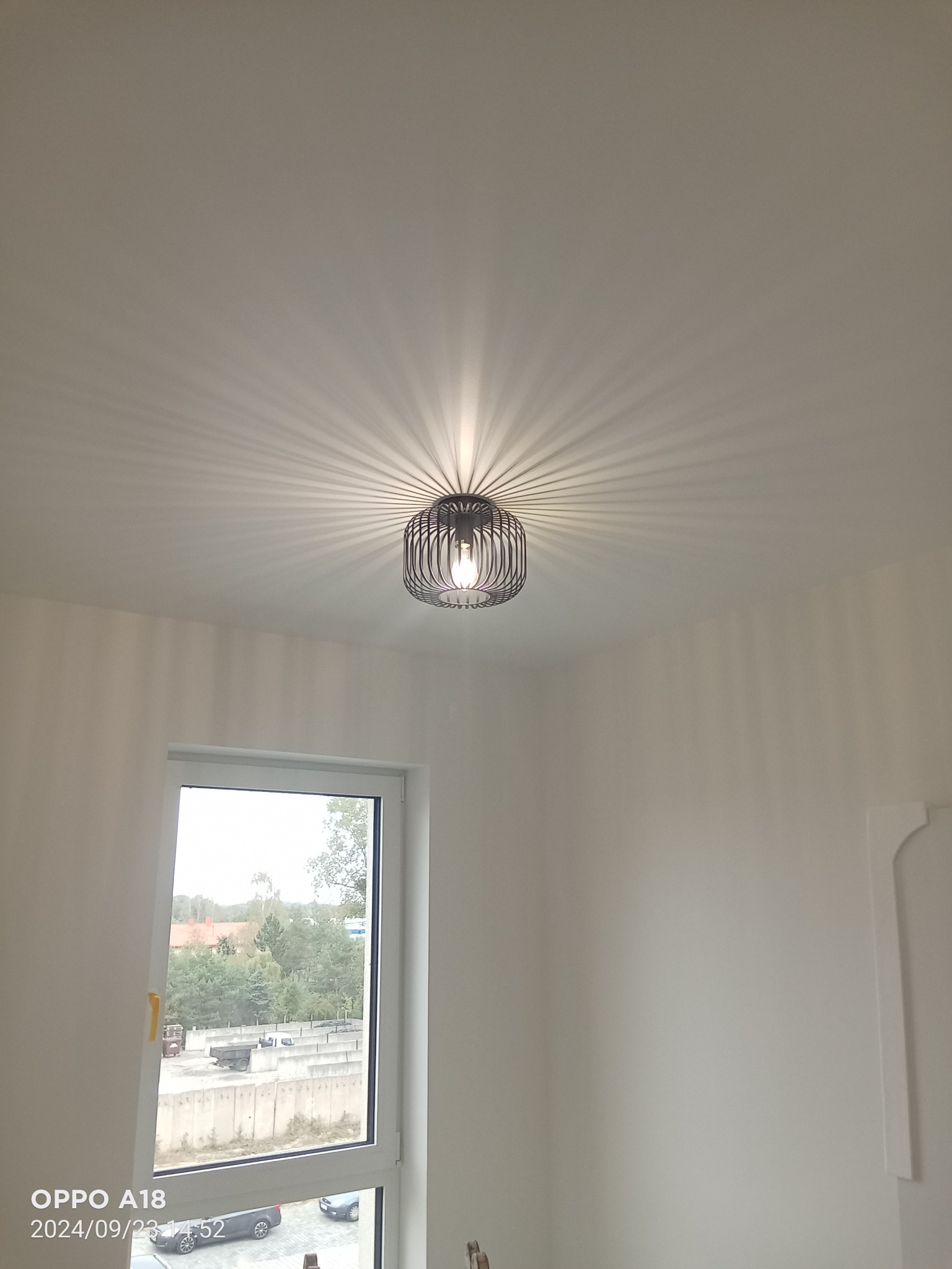 Nowoczesna lampa sufitowa z metalowym kloszem rzucająca promieniście światło na gładką, białą powierzchnię sufitu w nowym pomieszczeniu z oknem.