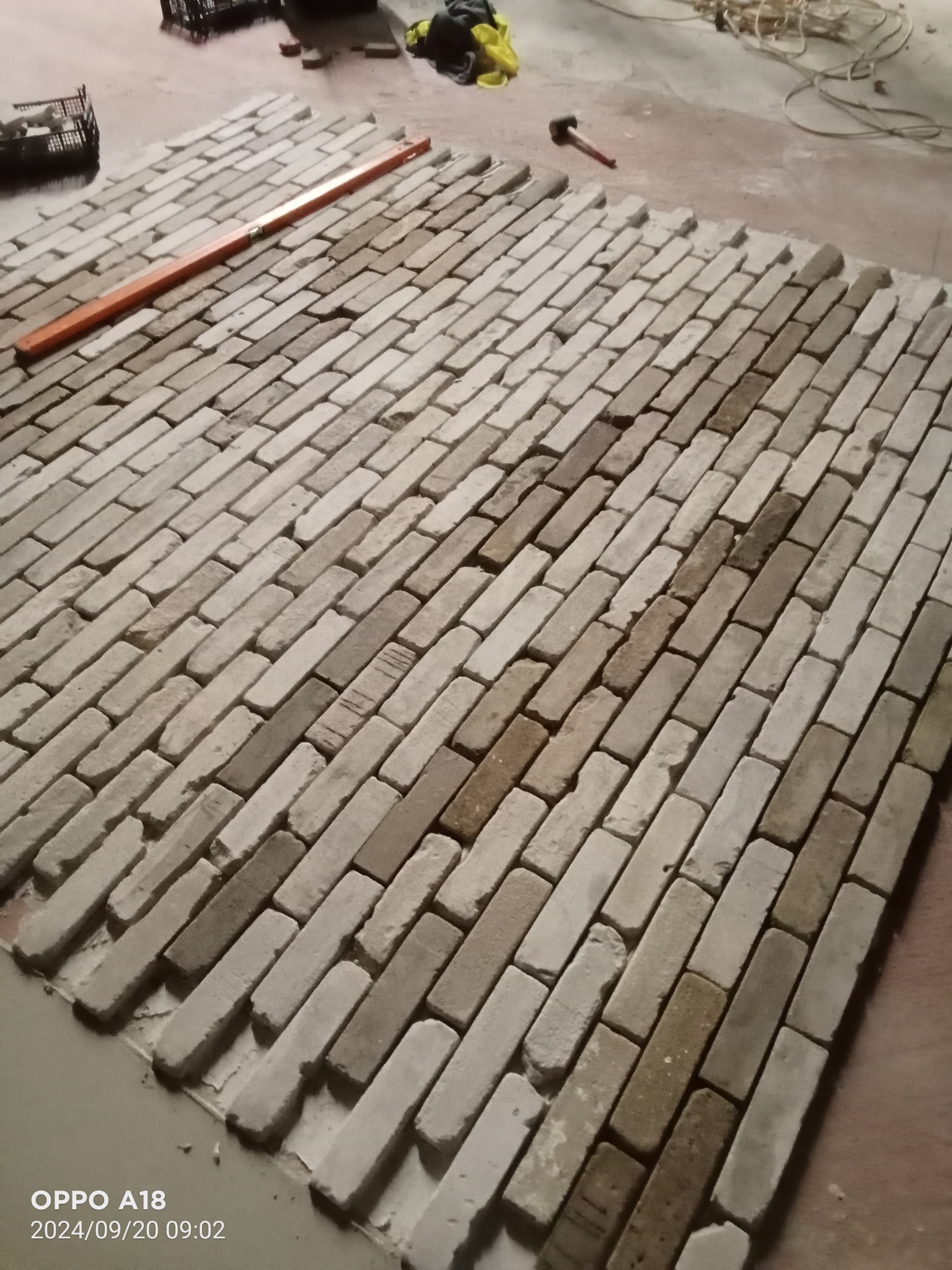 Układanie cegieł klinkierowych na podłodze, widoczna poziomica i młotek, etap prac wykończeniowych.