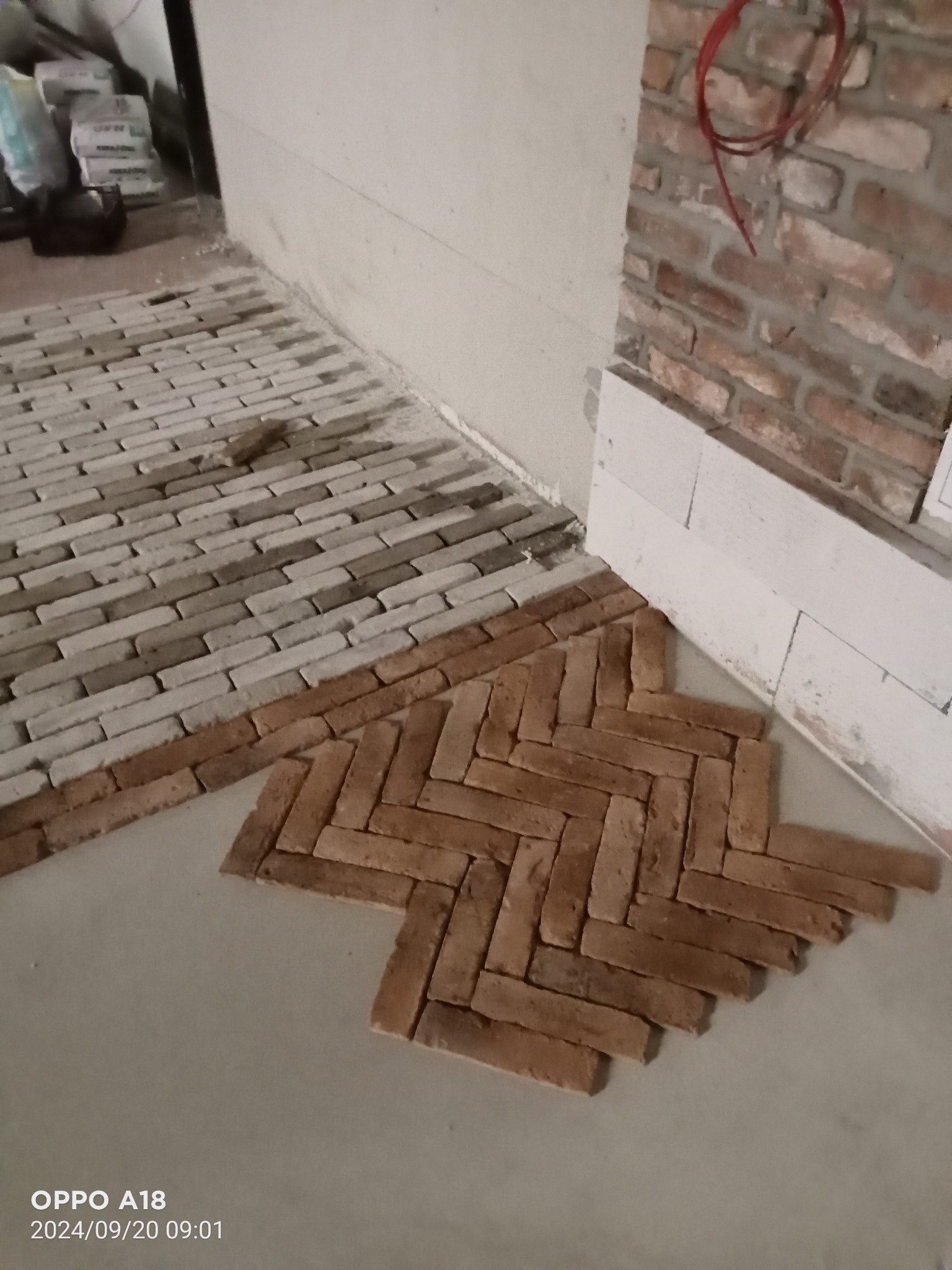 Fragment podłogi z cegieł ułożonych we wzór jodełki, widoczne także cegły ułożone rzędami wzdłuż ściany oraz worki z zaprawą w tle.