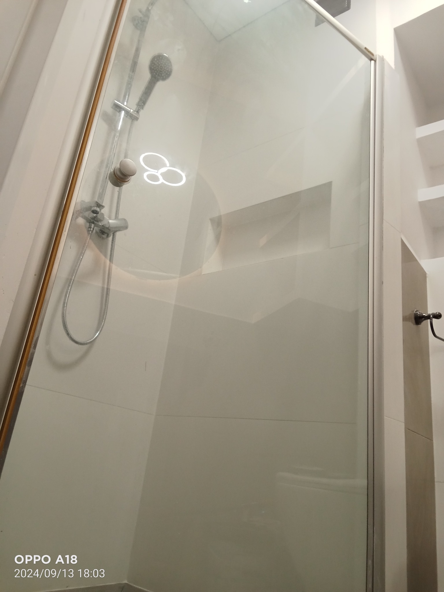 Szklana kabina prysznicowa z chromowaną armaturą, widoczna wnęka w ścianie wyłożonej białymi płytkami oraz półki w zabudowie, oświetlenie górne odbija się na szkle.