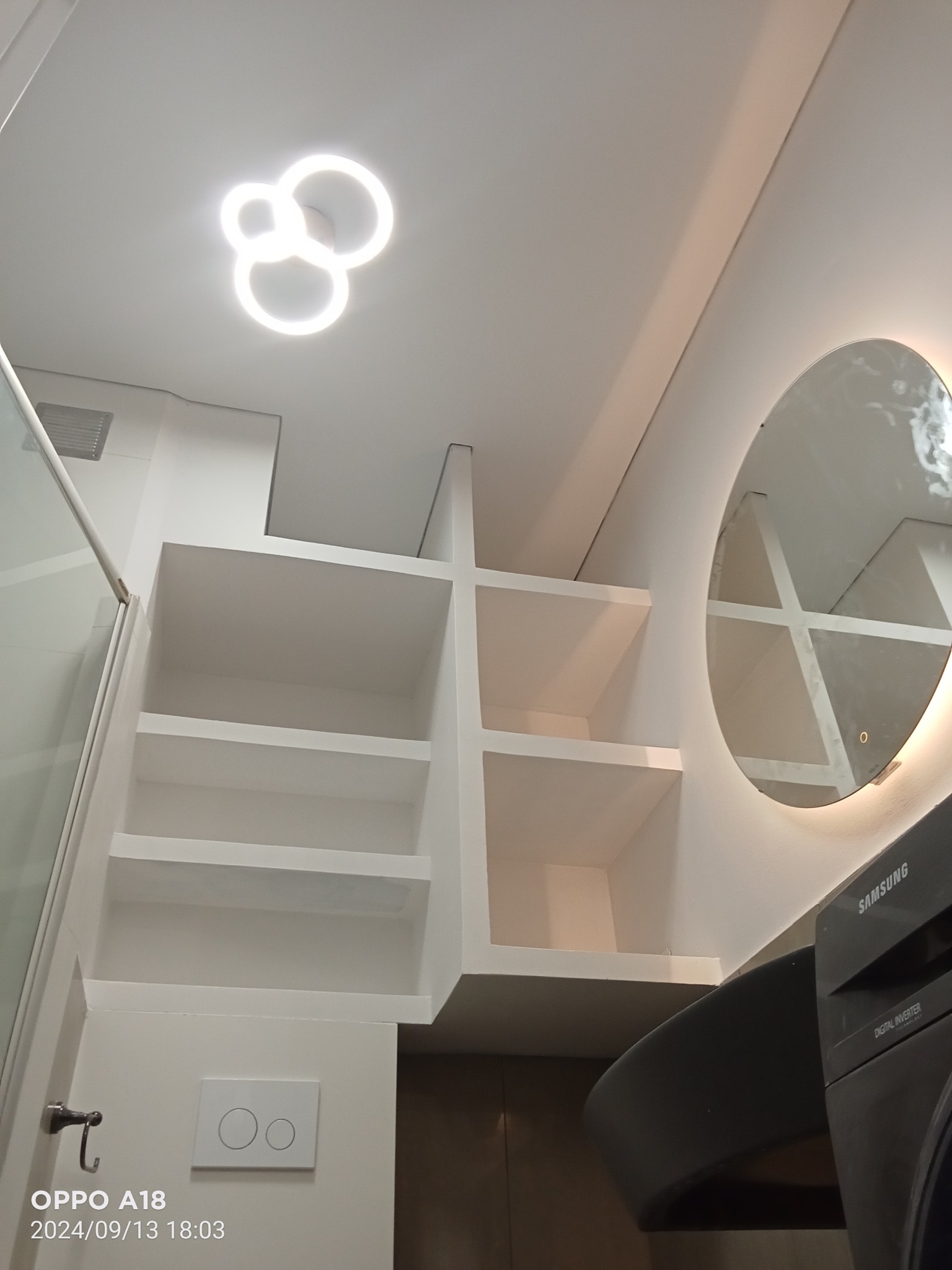 Nowoczesna łazienka z białą zabudową półek, okrągłym lustrem z podświetleniem LED i pralką Samsung, widok od dołu.